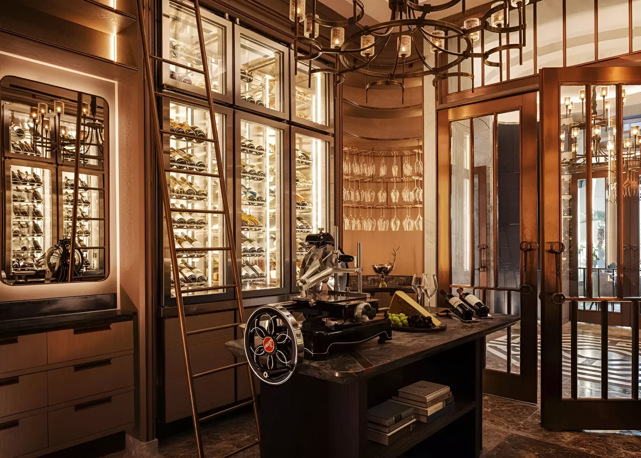 Bespoke wine room at La Veranda, One&Only Portonovi, Montenegro, featuring illuminated floor-to-ceiling wine displays, a central marble tasting table with vintage slicer, warm brass detailing, and layered ambient lighting designed to elevate the all-day dining experience.