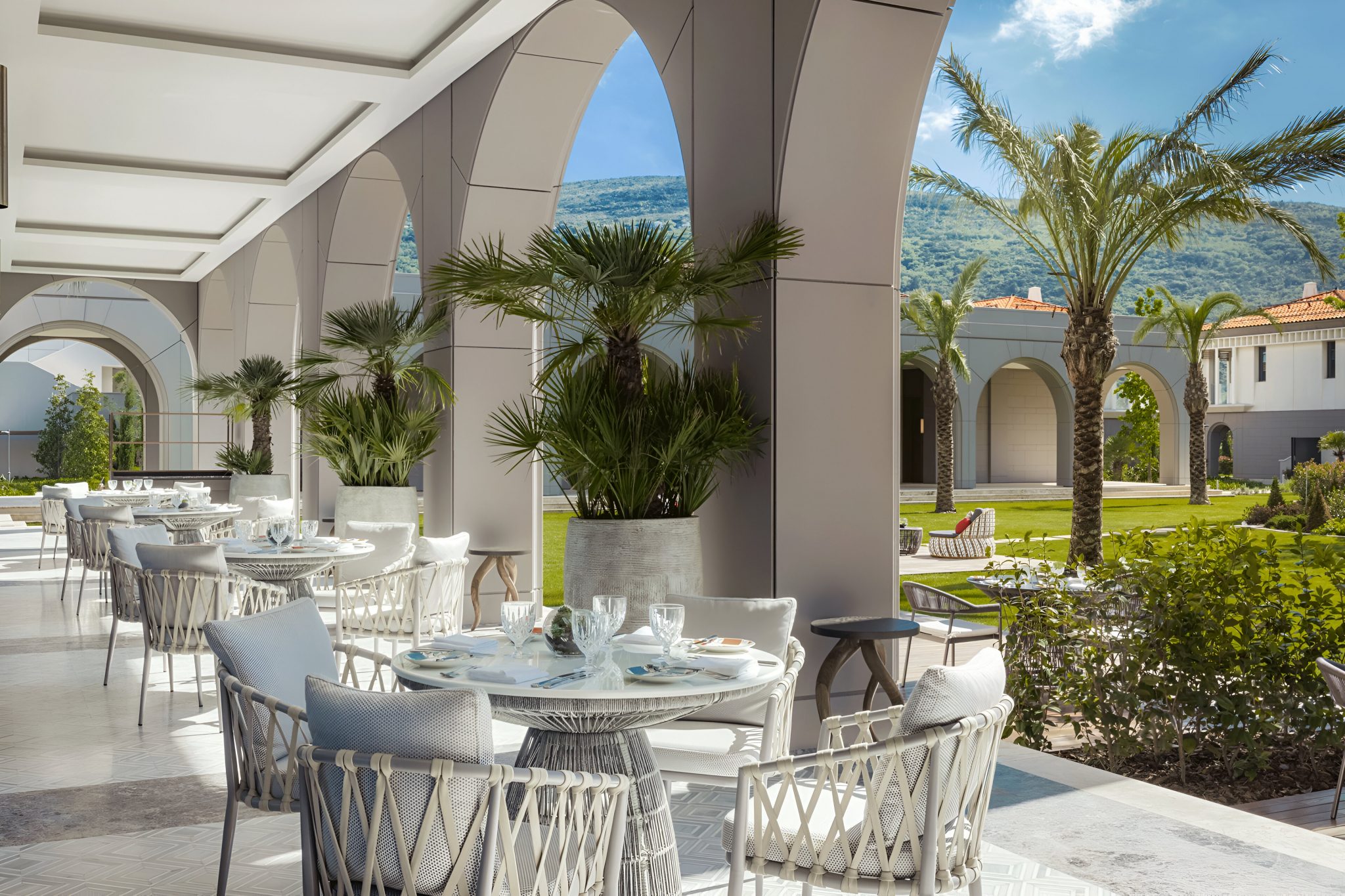 Outdoor dining terrace at La Veranda, the all-day dining restaurant at One&Only Portonovi, Montenegro, featuring arched colonnades, woven dining furniture, palm-lined gardens, and a light, Mediterranean-inspired setting overlooking the resort landscape.