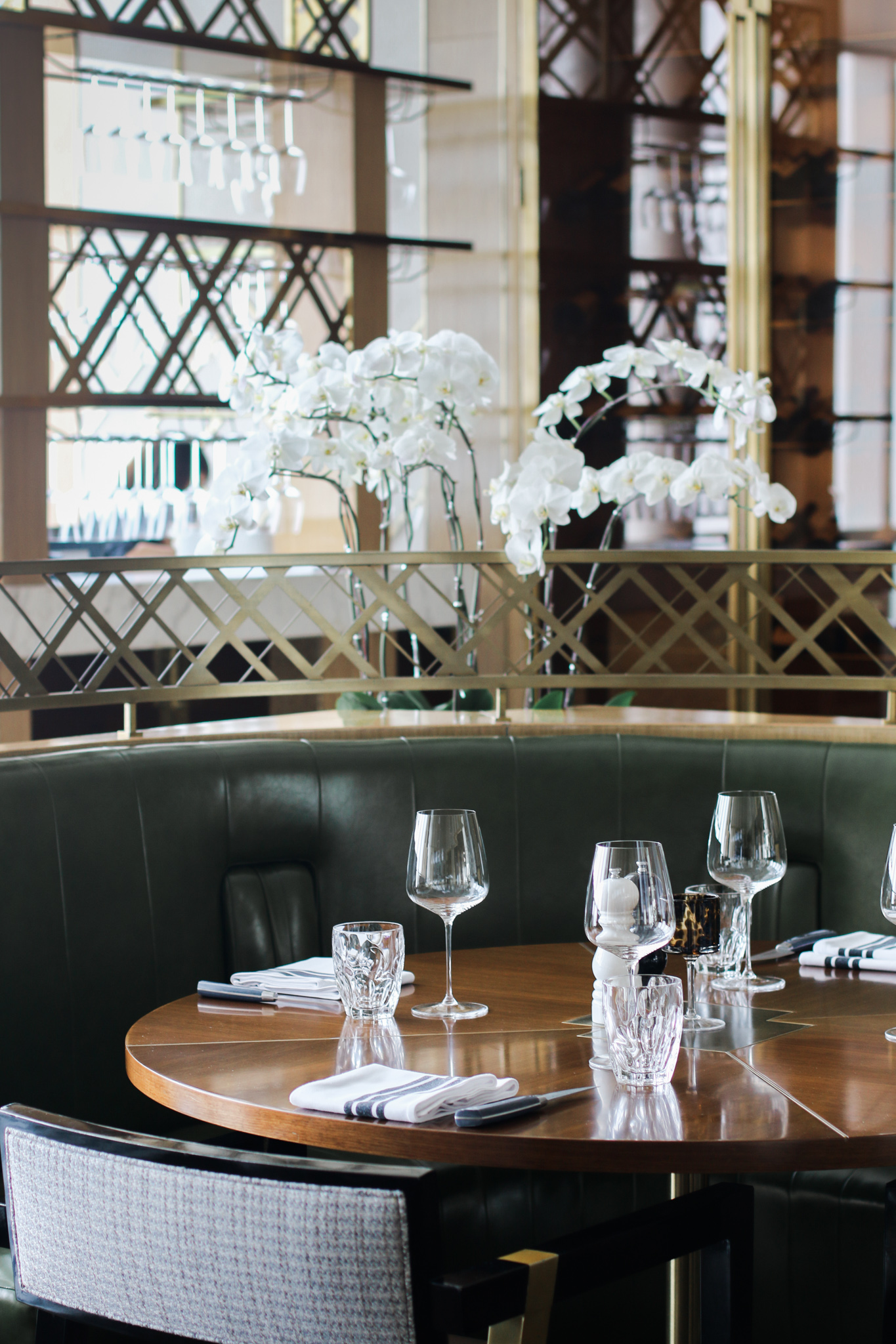 Table setting at Stellar Steakhouse featuring curved leather banquette seating, timber tabletop, and glassware framed by brass detailing and floral accents.