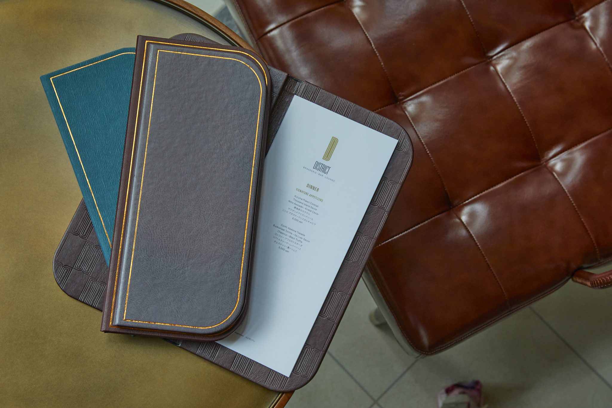Leather-bound menu design for The District, featuring a refined dark cover with gold edge detailing and textured inner panels, partially opened to reveal a minimalist dinner menu layout, styled on a leather chair to reflect the venue’s elevated brasserie-bar aesthetic and premium branding.