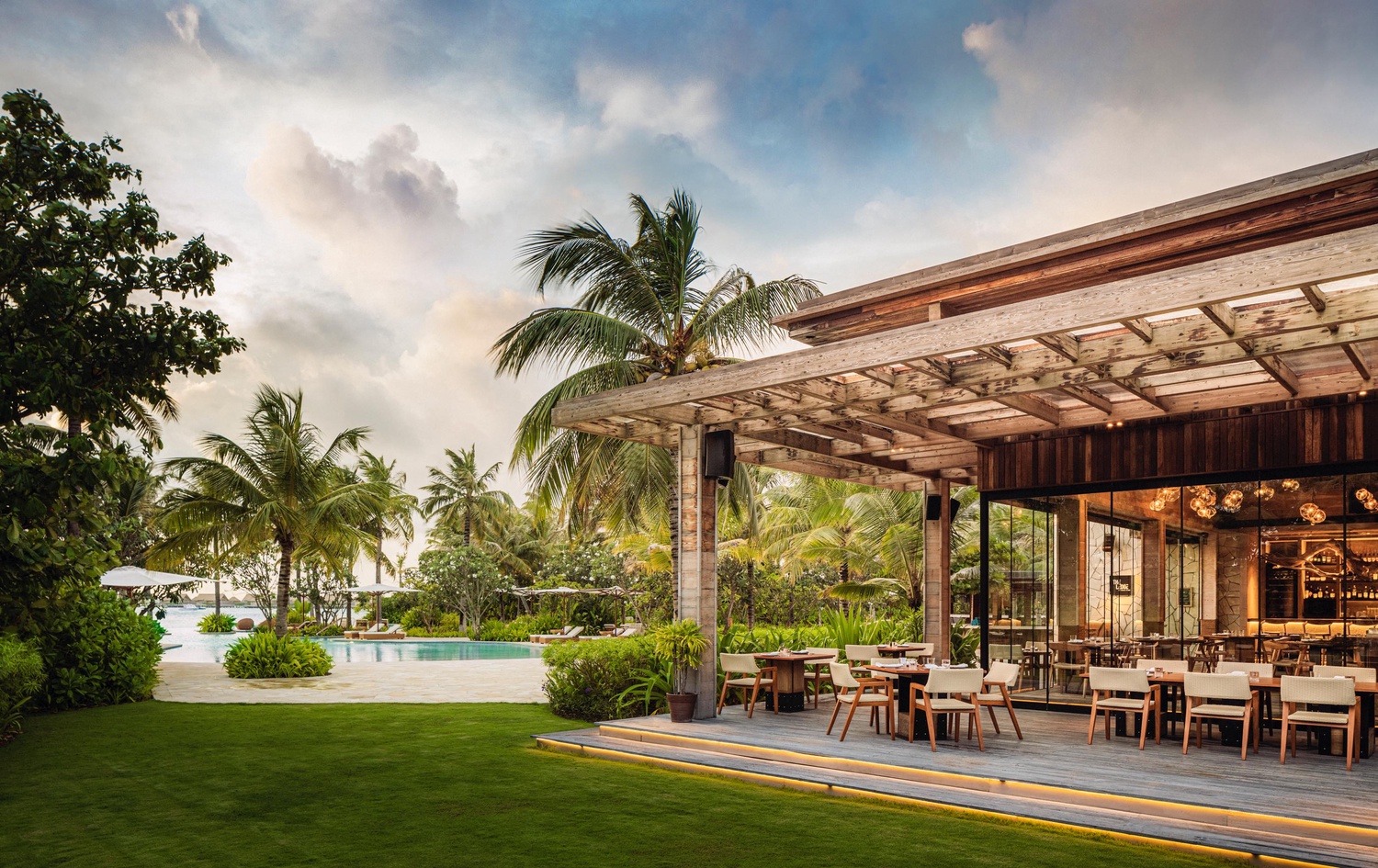Exterior view of The Ledge by Dave Pynt at Waldorf Astoria Maldives, featuring Social F+B’s interior design extending into an open-air dining pavilion with timber pergola, lush tropical landscaping, poolside setting, and warm evening lighting.