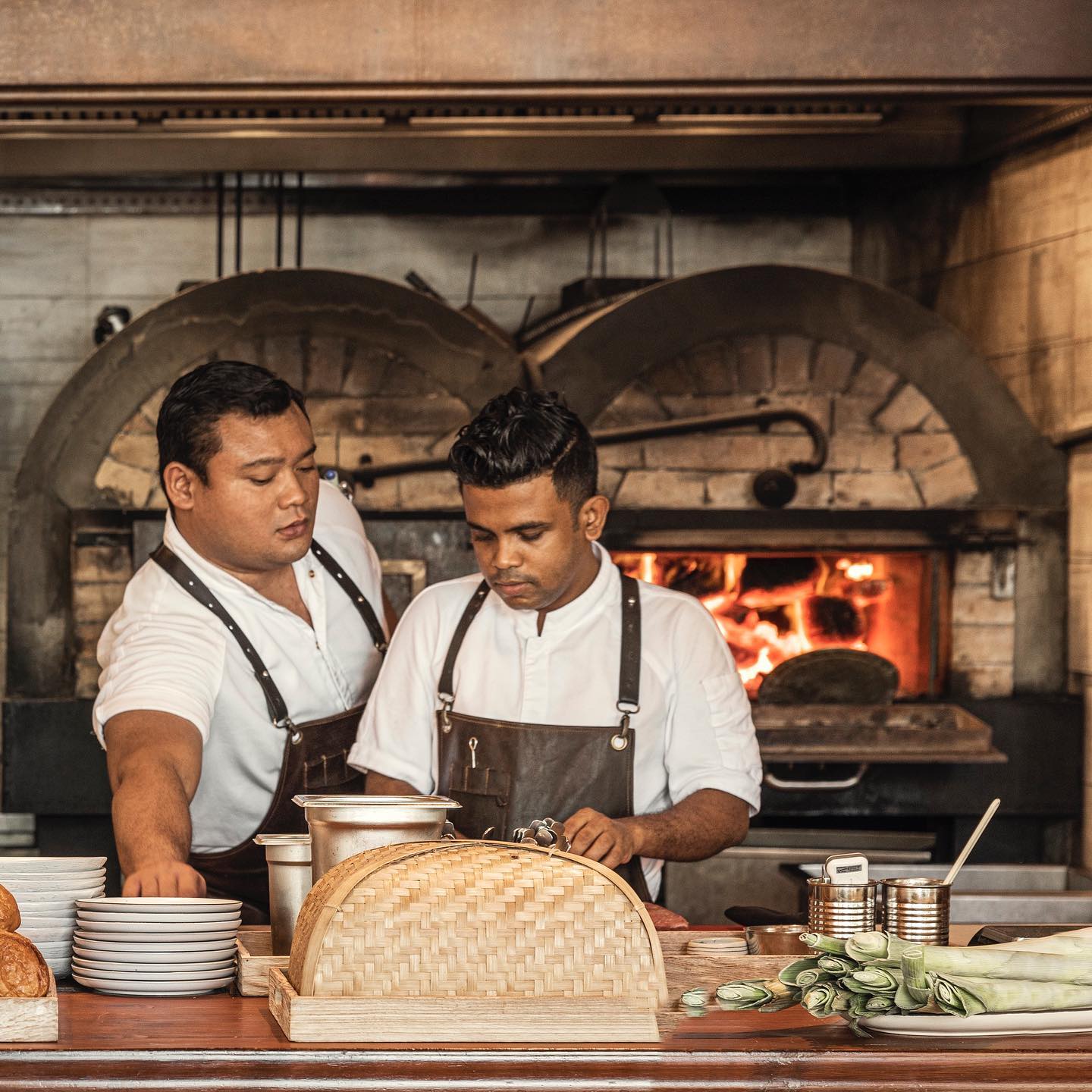 Chefs preparing dishes at The Ledge by Dave Pynt at Waldorf Astoria Maldives, working at an open kitchen with wood-fired oven, natural timber counters, and handcrafted details within an interior designed by Social F+B.