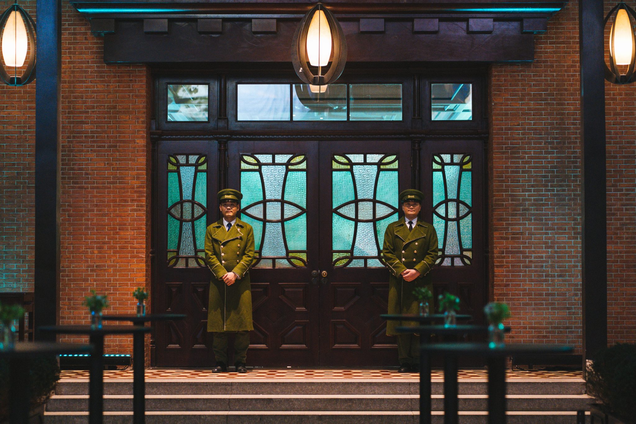Front entrance doors at Harrods Cha House Shanghai, featuring symmetrical stained-glass panels set within a red-brick façade, with two uniformed door attendants standing on either side under warm exterior lighting.