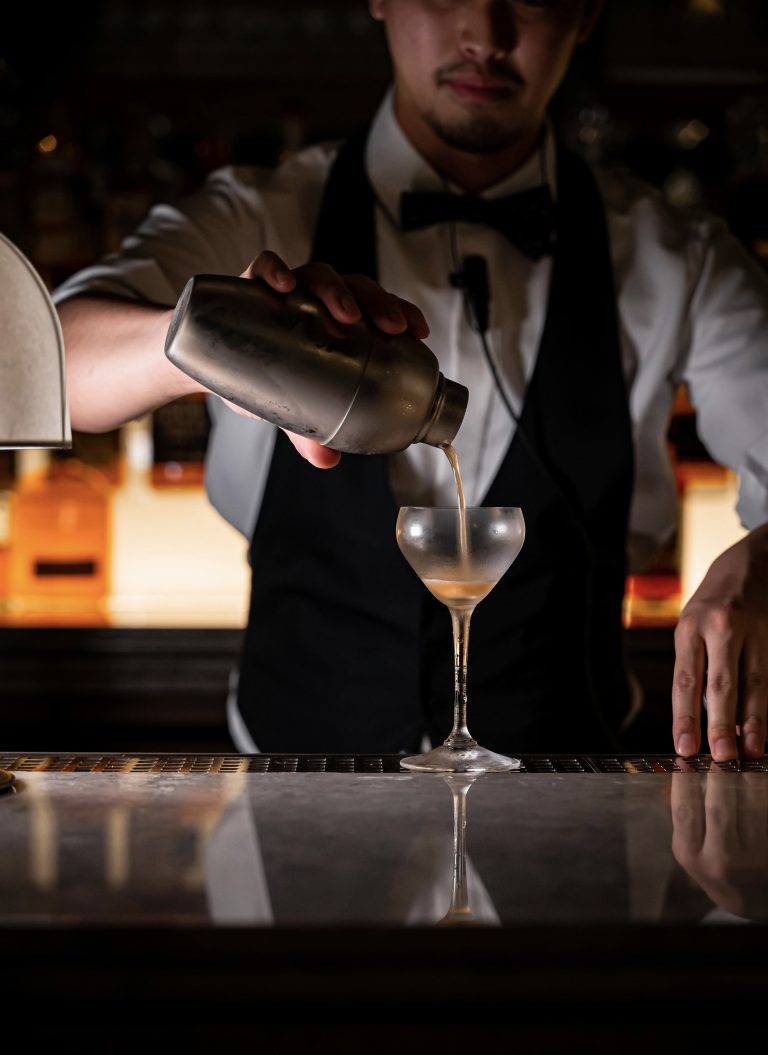 Bartender in formal attire pouring a cocktail from a metal shaker into a chilled stemmed glass at a dimly lit bar, capturing the precision, craft, and intimate atmosphere of an upscale cocktail experience