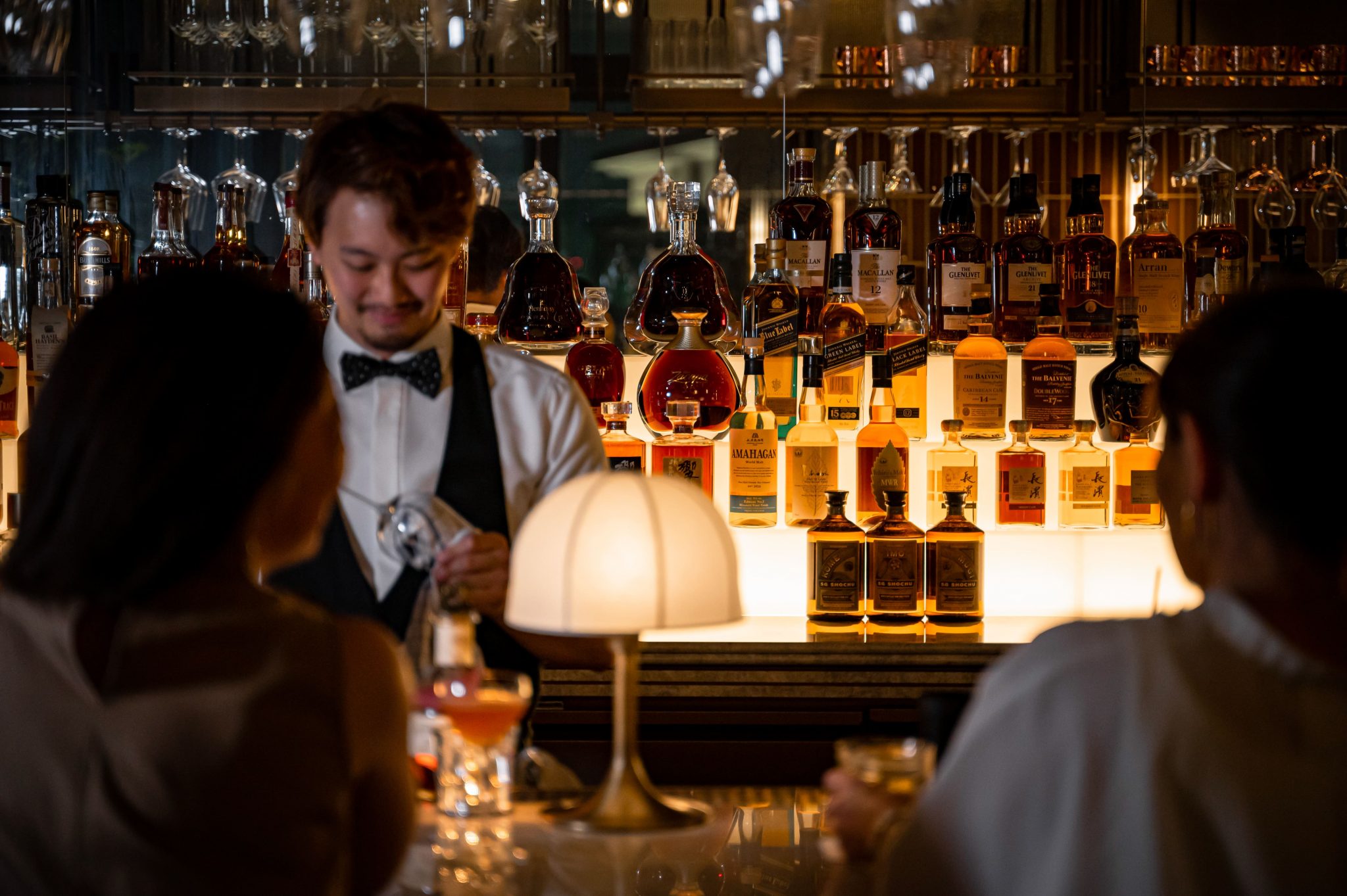 Intimate cocktail bar scene with a bartender preparing drinks in front of a curated back bar of premium spirits, softly lit glassware and guests seated at the counter, conveying a refined, evening-led hospitality atmosphere.