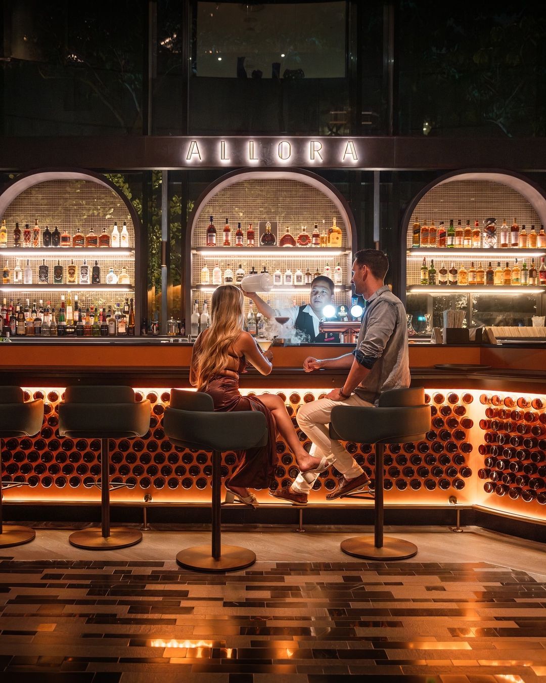 Evening bar scene at Allora Ristorante & Bar, Crowne Plaza Changi Airport, showing guests seated at the illuminated bar with arched back-bar shelving and warm amber lighting, capturing the refined, social atmosphere shaped through Social F+B’s concept and branding consultancy.