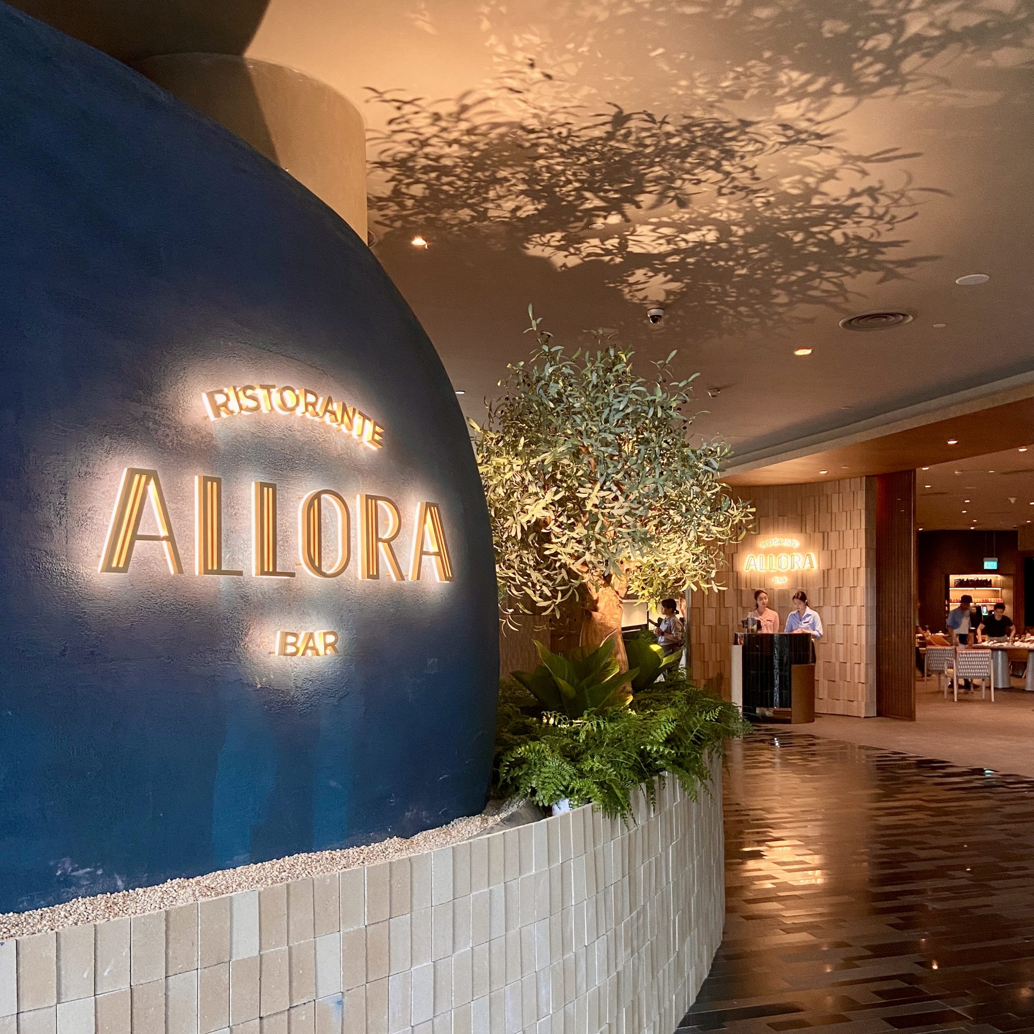 Illuminated Allora Ristorante & Bar signage integrated into a curved plaster wall and lush planting at Crowne Plaza Changi Airport, creating a warm, sculptural arrival moment shaped through Social F+B’s concept and branding consultancy.