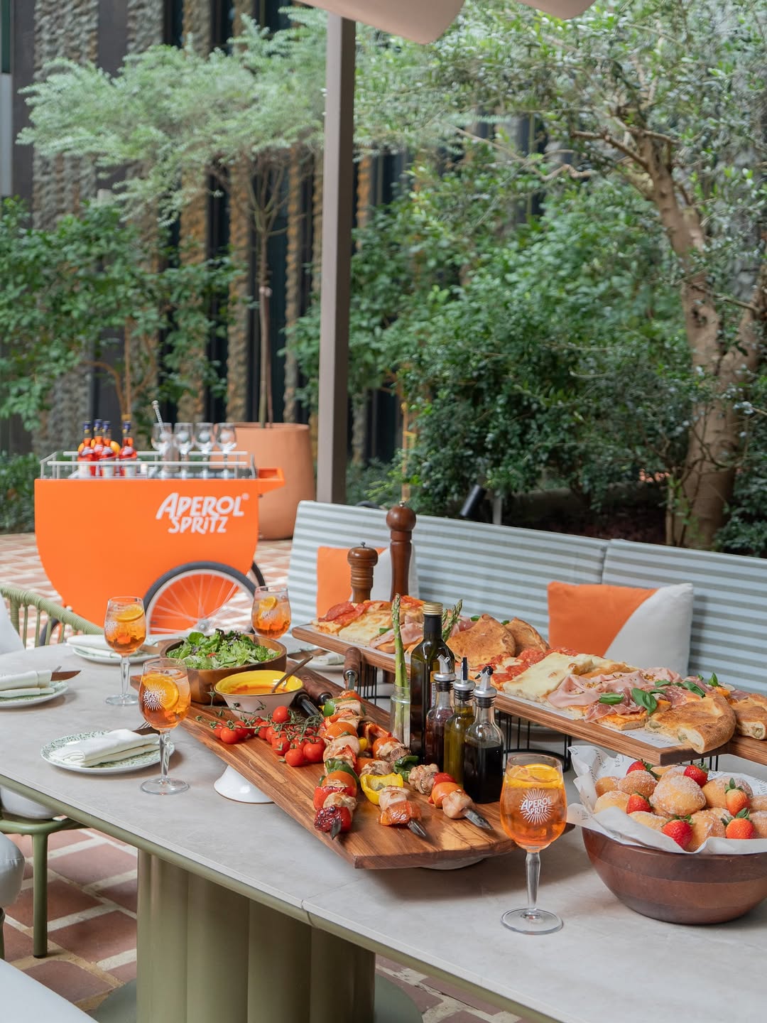 Outdoor terrace dining setup featuring an abundant Italian sharing spread at Allora Ristorante & Bar, with wooden boards of pizza, skewers, salads, bread, olive oil bottles, and Aperol Spritz cocktails served in branded glasses, styled in a lush garden setting to convey relaxed, social Mediterranean hospitality.