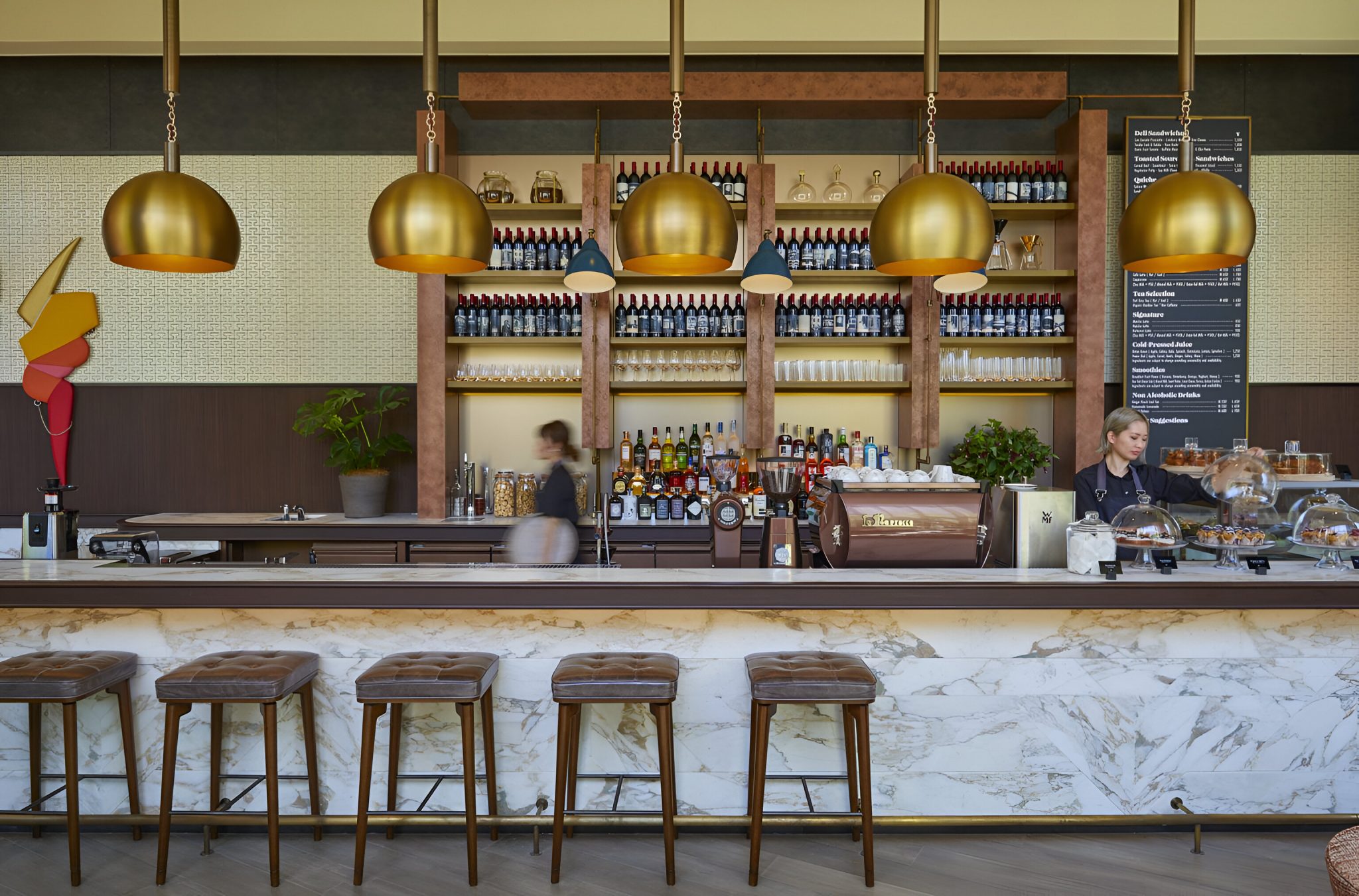 Coffee counter at The Jones, Kimpton Shinjuku Tokyo, featuring a marble bar front, brass pendant lights, back-bar shelving lined with bottles and glassware, and baristas in motion preparing drinks within a warm, neighbourhood café setting