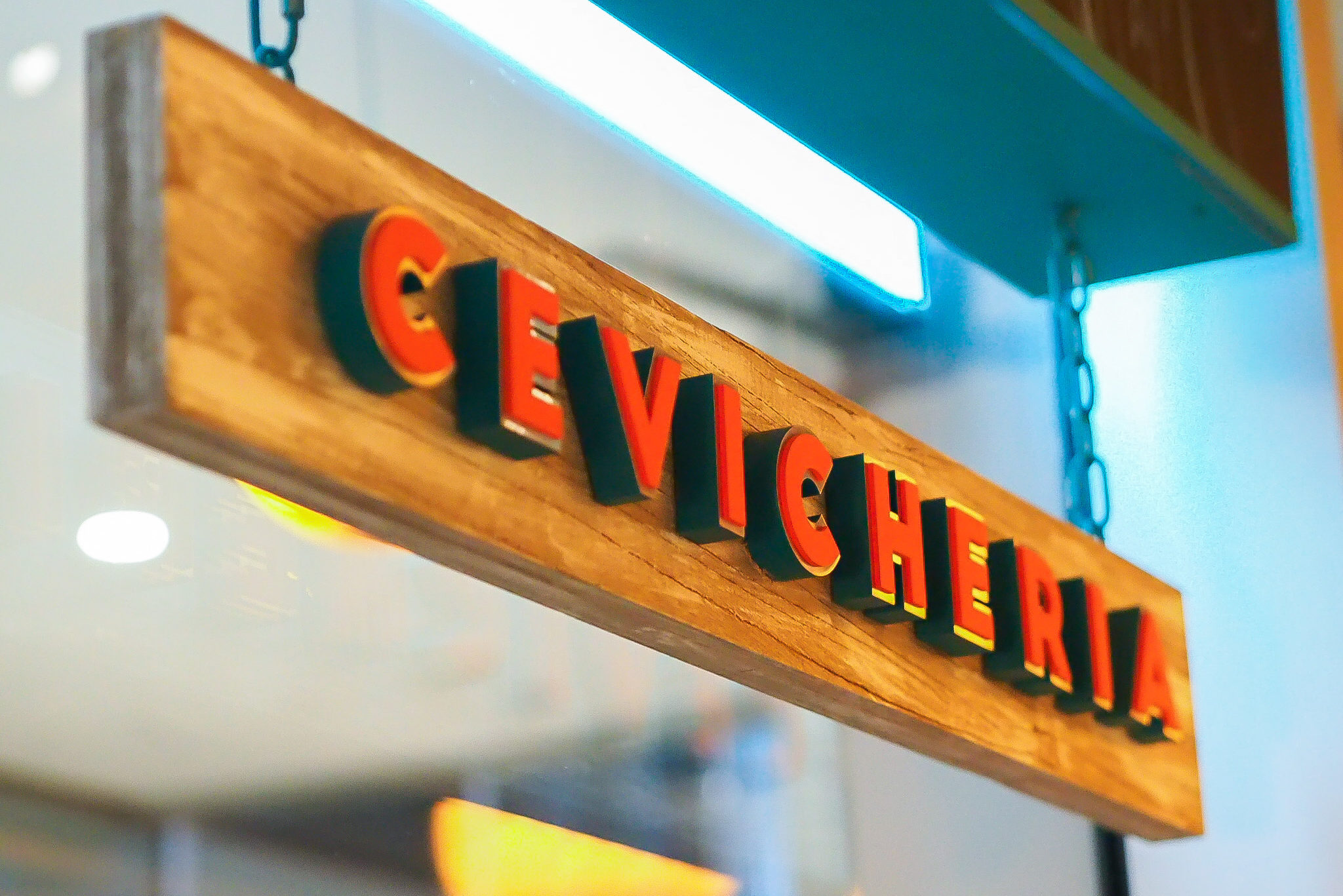 Hanging wooden banner signage for Tono Cevicheria, featuring bold three-dimensional red lettering mounted on a natural timber plank, suspended by metal chains and illuminated by warm ambient lighting as part of the restaurant’s branding and wayfinding design in Singapore.