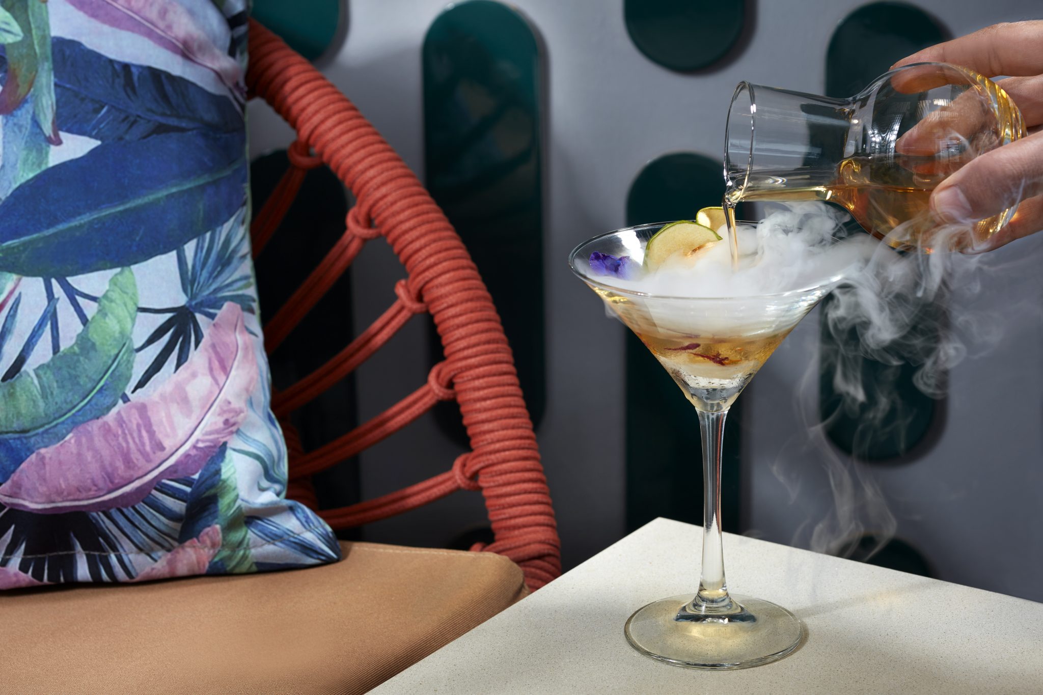 Close-up of a signature cocktail being poured tableside at Verandah Pool Bar & Grill, featuring a smoked martini glass with fruit garnish, set against tropical cushions and woven outdoor seating.