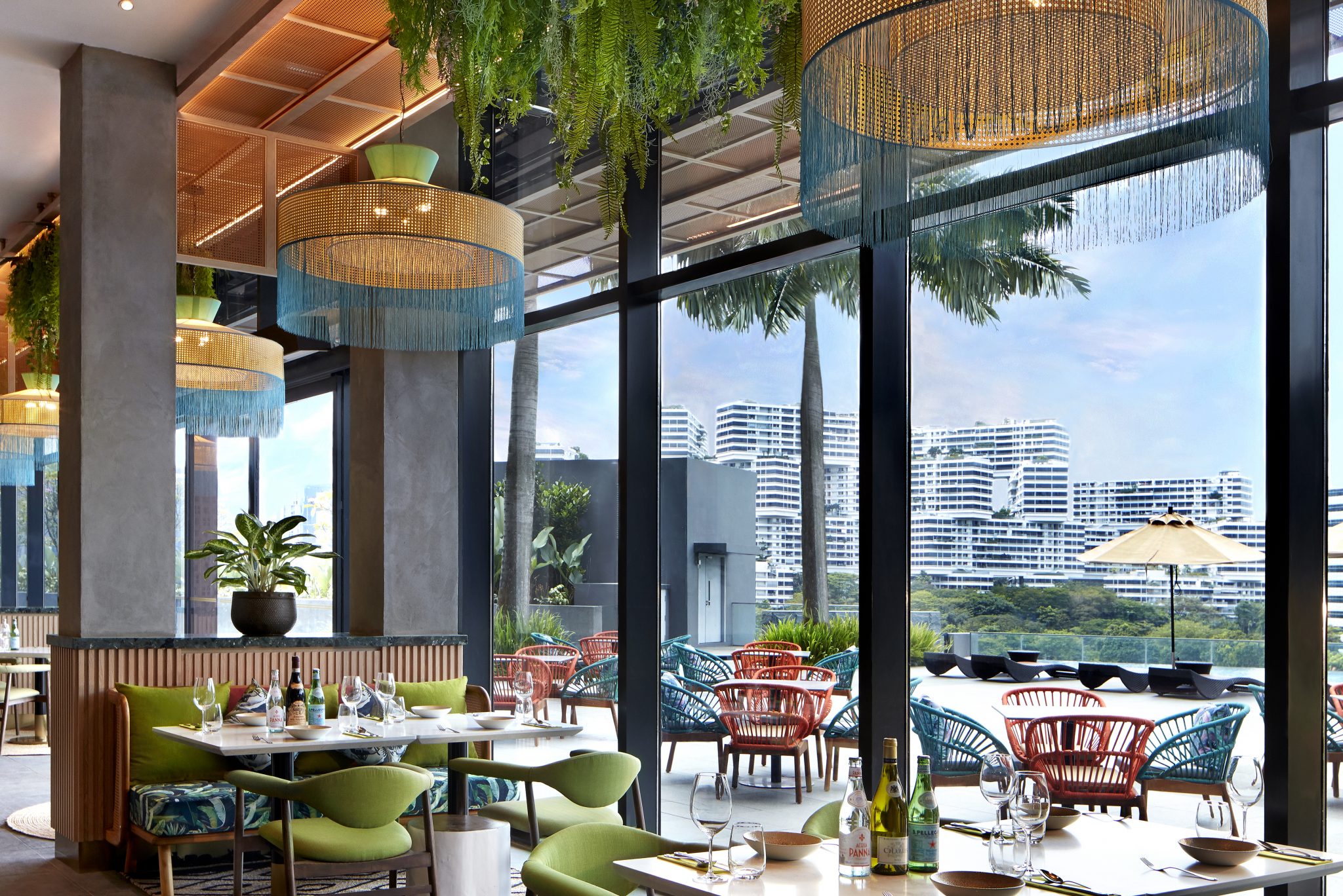 Verandah Pool Bar & Grill interior with floor-to-ceiling windows, tropical pendant lights, lush hanging greenery, and indoor-outdoor seating overlooking the pool and city skyline.